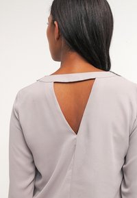 Lichtgrijze blouse met een diepe V-rugontwerp, voorzien van een sleutelgatopening, lange mouwen en een subtiele glans in de stoftextuur.