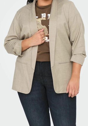 Osoba ubrana w beżową marynarkę na brązowym t-shircie z nadrukiem lamparta, zestawione z ciemnoniebieskimi dżinsami. Jedna ręka trzyma klapę marynarki.