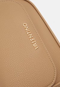 Beige leren tas met een textuur en een gladde, gebogen rand. Bevat een gouden, geëmbosserd "VALENTINO" logo aan de zijkant. Eenvoudig ontwerp met minimale hardware.
