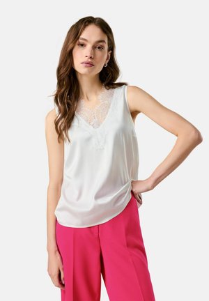 Frau mit langen braunen Haaren, die ein weißes ärmelloses Oberteil mit Spitzenbesatz und leuchtend pinke Hosen trägt, posiert mit einer Hand in der Hüfte.