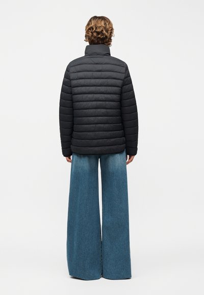 Άτομο με κοντά κυματιστά μαλλιά που φοράει μαύρο καπιτονέ μπουφάν και φαρδιά μπλε τζιν παντελόνια, στέκεται γυρισμένο με την πλάτη σε λευκό φόντο.