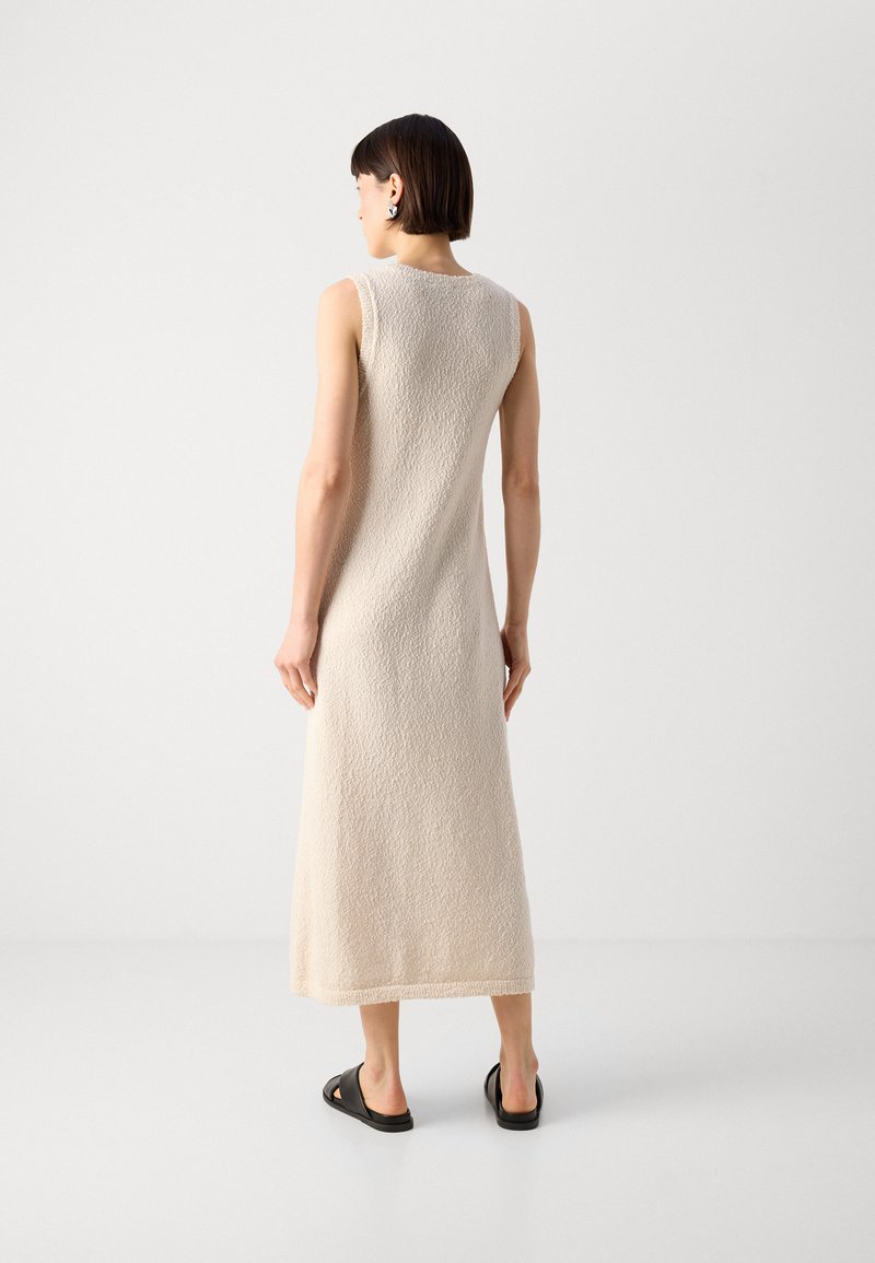 Robe maxi sans manches beige avec une surface texturée, dotée d'une coupe ample et d'une fente latérale, assortie de sandales noires.