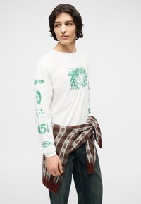 Νεαρός άνδρας με σκούρα μαλλιά φοράει λευκό μακρυμάνικο μπλουζάκι με σχέδια και τζιν, με πουκάμισο καρό δεμένο γύρω από τη μέση του, κοιτάζοντας προς τα αριστερά.