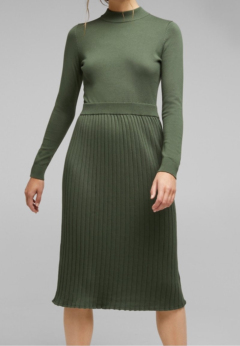 Groene geribbelde gebreide jurk met lange mouwen, een hoge halslijn en een strakke bodice die overgaat in een uitlopende geplooide rok. Gladde textuur.