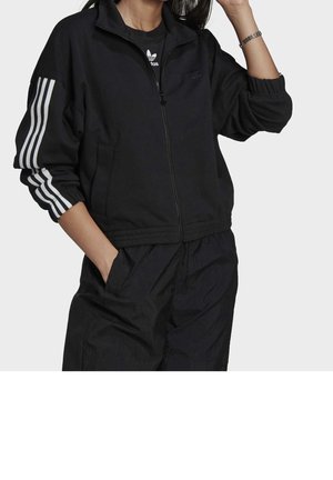 Kvinde i sort Adidas træningsjakke med tre hvide striber på ærmerne, hænderne i lommerne, iført matchende sorte bukser og armbånd.