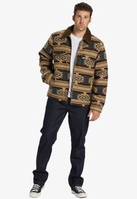 Brown patterned jacket with a faux fur collar, featuring geometric designs in beige, blue, and brown; paired with dark denim jeans and sneakers.