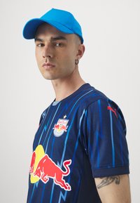 Cappello blu, maglia blu scuro con motivo a onde, con logo Red Bull e cerchio giallo. Maniche corte e dettagli del logo sulle maniche.