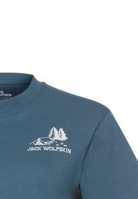 T-shirt in cotone blu con collo rotondo e maniche corte, decorata con una grafica bianca di alberi e una tenda, e il testo "JACK WOLFSKIN".