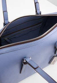 Niebieska skórzana torebka z lawendy z dwoma uchwytami, otwartym zamkiem na górze, wewnętrzną kieszenią na zamek oraz markową metalową zawieszką z przodu.