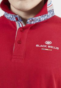 T-shirt polo rouge avec un col à motifs bleu et blanc, doté de deux boutons et portant "BLACK WELLIS" brodé sur la poitrine.