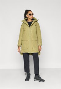 Olijfgroene parka met capuchon, twee zakken aan de voorkant en ritssluitingdetails. Gecombineerd met zwarte broek en veterschoenen. Soepele textuur, casual ontwerp.