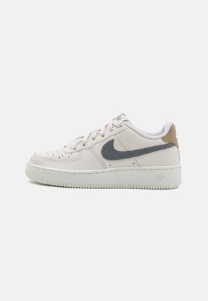 Zapatilla blanca Nike Air Force 1 con logo swoosh gris y lengüeta del talón beige mostrada de perfil sobre un fondo liso.
