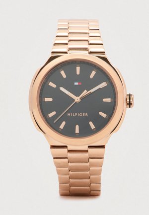 Rózsaarany színű fém karóra fekete számlappal, arany órajelzőkkel és mutatókkal, valamint egy kis Tommy Hilfiger logóval a 12 órai pozíció alatt.