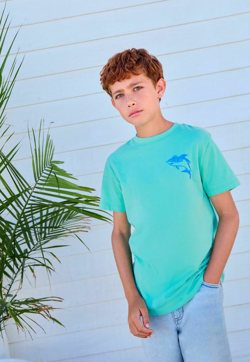 Garçon aux cheveux roux bouclés portant un t-shirt turquoise avec un motif requin bleu et un jean bleu clair, debout à côté d'une plante verte.
