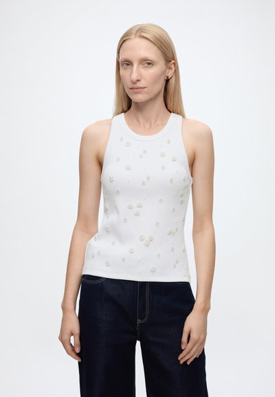 Femme aux cheveux blonds et raides portant un haut blanc sans manches orné de petites fleurs et un pantalon bleu foncé, debout sur un fond blanc.
