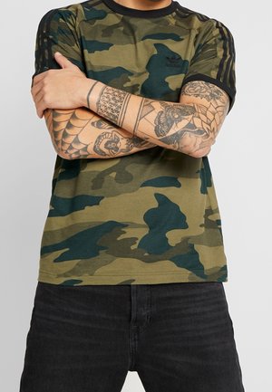Man met gekruiste armen, gekleed in een groen-zwart camouflage Adidas-shirt en zwarte broek, met gedetailleerde tatoeages op beide onderarmen.