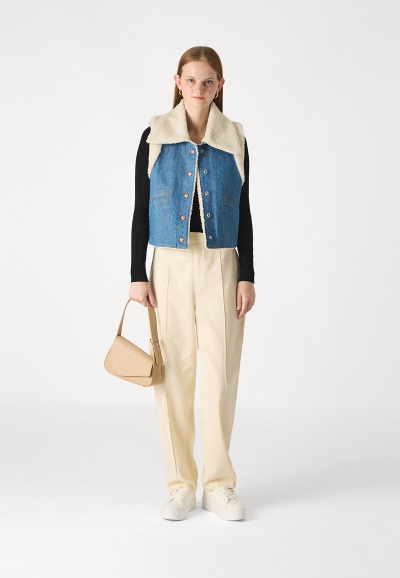 Jeansowa kamizelka z kołnierzem z wełny owczej, miedziane guziki, w połączeniu z kremowymi spodniami o szerokich nogawkach i beżową torebką. Białe sneakersy dopełniają całość.