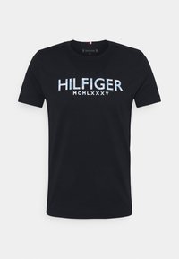 Mustat lyhythihaiset T-paidat, joissa on rintaosassa sinisellä ja valkoisella painettuna "HILFIGER MCMLXXXV".