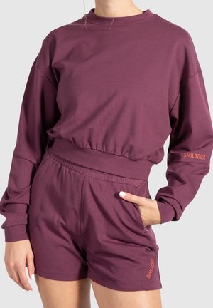 Persoon die een bijpassende paarse top met lange mouwen en korte broek draagt met de tekst "SMILODOX" op de mouw en korte broek, één hand in de zak.