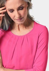 Fuchsia Bluse mit 3/4-Ärmeln, rundem Ausschnitt und plissierten Details an der Vorderseite. Weicher, glatter Stoff mit einem leichten Glanz.