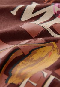 Brown cotton fabric featuring a graphic print in orange, pink, and cream. The design includes abstract shapes and textured details.