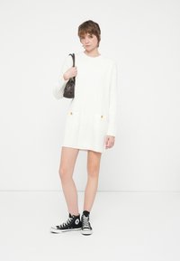 Tânără îmbrăcată într-o rochie albă din tricot cu model torsadă, cu buzunare, pantofi sport negri cu glezna și purtând o mică geantă de umăr neagră.