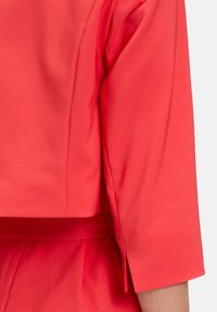 Blusa di tessuto corallo con maniche a tre quarti, caratterizzata da una texture morbida e una vita con cintura. Dettagli delle cuciture piatte lungo le maniche.