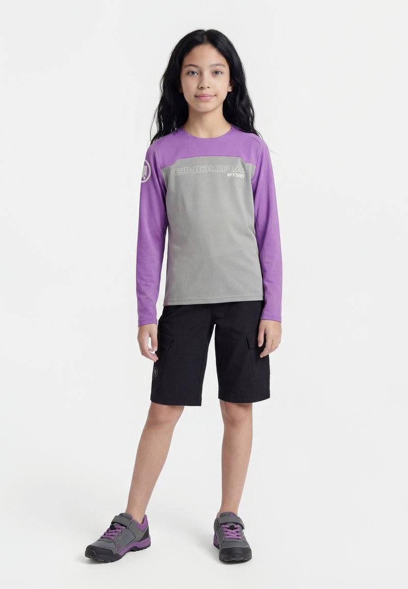 Persona joven de pie, vistiendo una camiseta de manga larga gris y púrpura Endura MT500, pantalones cortos cargo negros y zapatillas gris púrpura sobre fondo blanco.