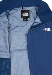 Blå jacka med nätfoder, som har en etikett från The North Face och en dragkedjeficka på höger sida.