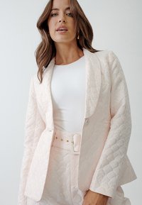 Quilted pink blazer featuring a notched lapel, single button closure, and 3/4 sleeves, paired with a fitted white top.