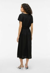 Black midi dress with short sleeves, elastic waist, flowing fabric, and a smooth texture. Silver heeled sandals complete the look.