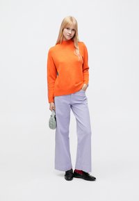 Oranžinis megztinis su aukštu kaklu, derinamas su platėjančiomis levandų spalvos dryžuotomis kelnėmis. Aksesuarai: maža žalia rankinė ir juodi batai.