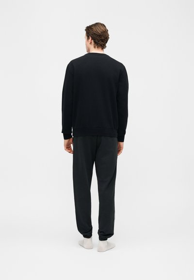 Άνδρας που στέκεται με την πλάτη στον θεατή, φορώντας μαύρο φούτερ, μαύρο αθλητικό παντελόνι και λευκές κάλτσες, πάνω σε απλό λευκό φόντο.