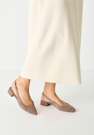 Voeten in taupe suede slingback schoenen met een lage hak, gecombineerd met crèmekleurige wijde broek tegen een eenvoudige lichte achtergrond.