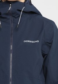 Veste Didriksons bleu marine avec fermeture éclair à l'avant et capuche, cordons blancs, portée par une personne sur un fond clair uni.
