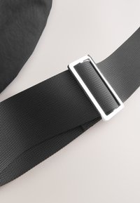 Black woven adjustable strap threaded through a rectangular silver metal buckle on a light background.