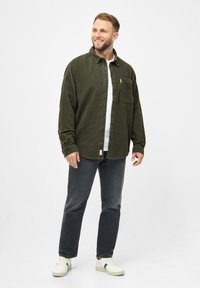 Chemise en velours côtelé vert olive avec des boutons sur le devant, une poche poitrinaire unique et un col, portée par-dessus un T-shirt blanc, associée à un jean sombre et des baskets blanches.