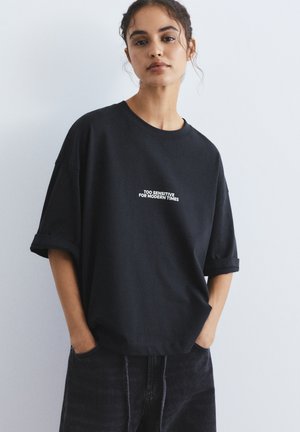 Mlada ženska z kodrastimi lasmi nosi ohlapno črno majico s kratkimi rokavi, zavihanimi na roke, z belim napisom "PREVEČ OBČUTLJIVA ZA SODOBNE ČASE", roke v žepih.