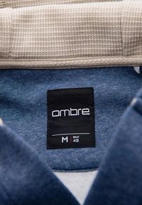 Tejido azul oscuro con una textura suave, con una etiqueta "ombre" negra impresa en blanco. El cuello es de color crema y texturizado. Talla M, EUR 48.