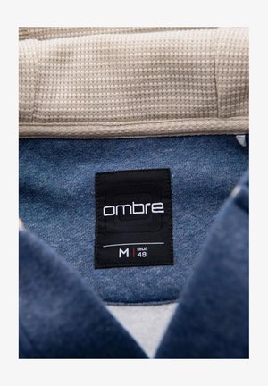 Dark blue fabric with a soft texture, featuring a black "ombre" label printed in white. The collar is cream-colored and textured. Size M, EUR 48.