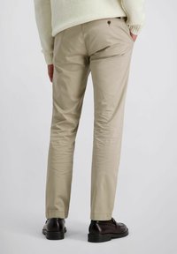 Man in beige broek, crèmekleurige trui en donkerbruine nette schoenen, staande met één hand in zijn zak tegen een effen achtergrond.