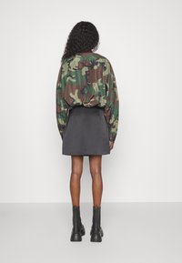 Donna con capelli ricci che indossa una camicia a maniche lunghe in camouflage, una gonna nera a trapezio e stivali neri chunky alla caviglia, in piedi di spalle.