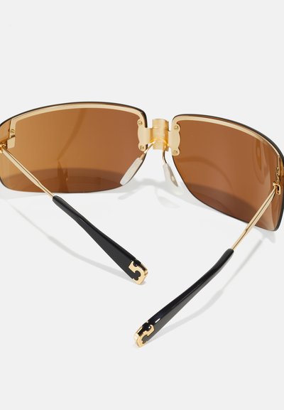 Lunettes de soleil oversize teintées de brun avec une monture en métal doré, présentant des branches en plastique noir et des accents dorés aux extrémités des tempes.