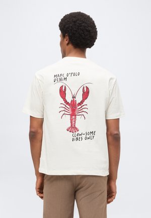 Hombre con camiseta blanca con gráfico de langosta roja y texto "Marc O'Polo Denim" y "Claw-some vibes only" en la espalda.