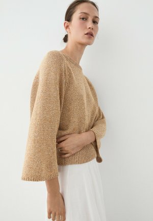 Femme aux cheveux attachés portant un pull beige ample et texturé avec de larges manches et une jupe blanche fluide sur un fond uni.