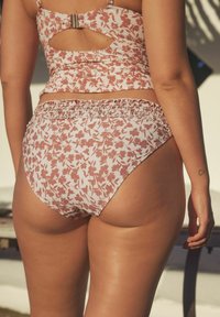 Cvečni bikini v mehki roza barvi z nagubanem pasom. Ima izrez na hrbtu in nastavljive naramnice. Gladka tekstura z nežnim vzorcem.