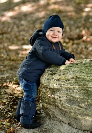 Sininen toppatakki, jossa on ruskeita yksityiskohtia, tumma sininen hattu, farkunsiniset farkut ja mustat saappaat. Lapsi seisoo luonnonläheisen, karheatuksen kalliolla.