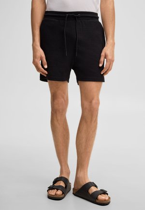 Mężczyzna w czarnych szortach ze sznurkiem i czarnych sandałach na dwóch paskach stoi na szarym tle, ręce swobodnie opuszczone wzdłuż ciała.