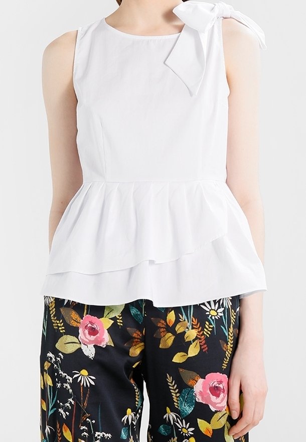 Femme portant un haut blanc sans manches avec un nœud à l'épaule et un ourlet à volants, associé à un pantalon noir avec imprimé floral coloré.