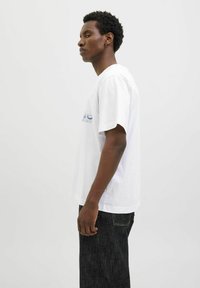 Weißes Baumwoll-T-Shirt mit kurzen Ärmeln, mit einem blauen Druckdesign auf der Vorderseite. Das Modell trägt dunkle Jeans und steht im Profil.